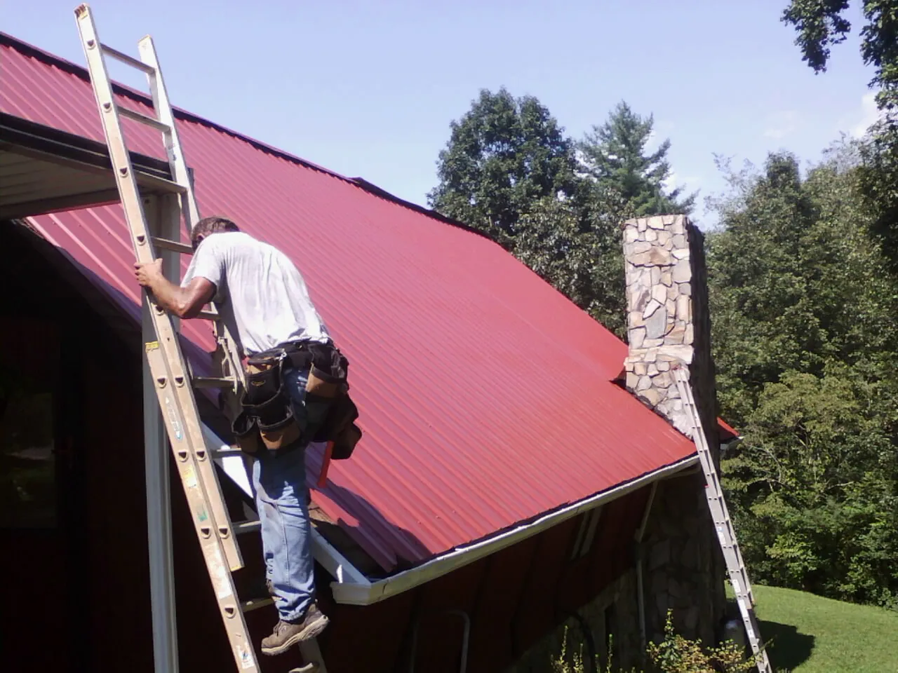 Roofer on a ladder beside a red metal roof and stone chimney — Soffit & Fascia Repair by Roofing Brothers in Junction City